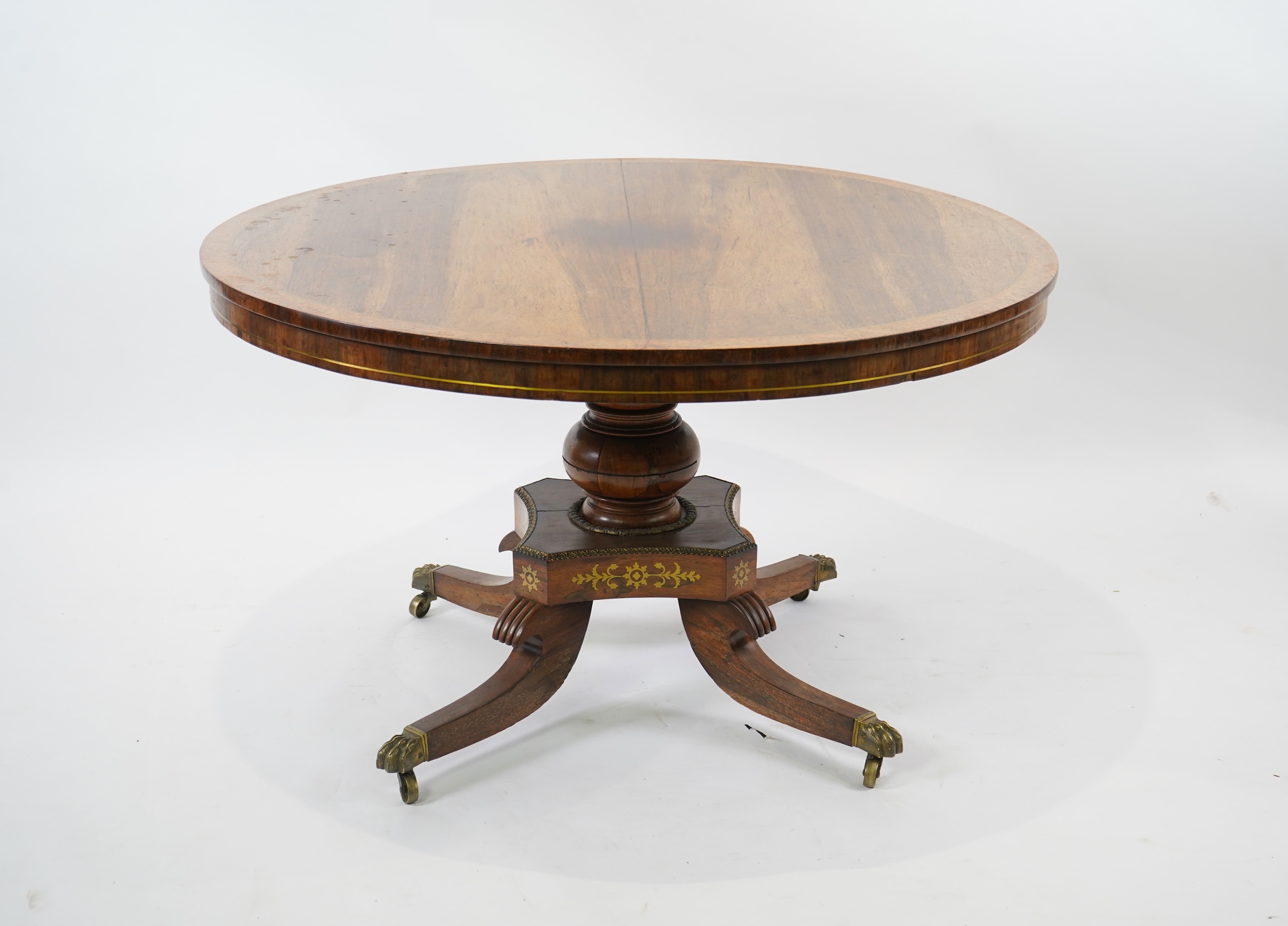 A Regency cut brass inlaid and gilt metal mounted circular rosewood breakfast table, the bird's-eye maple banded tilt top on baluster column, concave platform and quadruple downswept base with cast claw caps and castors,
